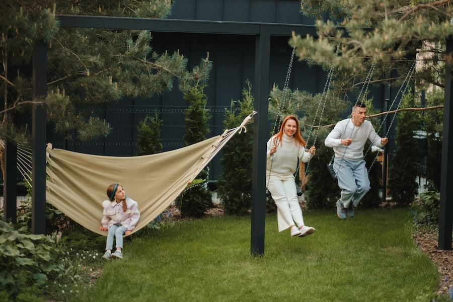 a family spenidng time outdoors, in their green backyard. Parents on the swing, daughter in a hammock